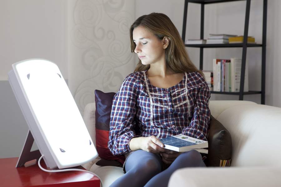  A woman sits on a couch and reads a book with a bright light therapy lamp next to her.